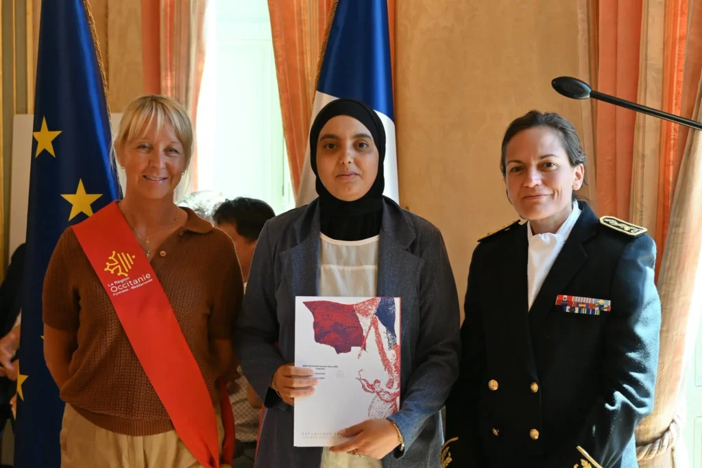 Photo de cérémonie de naturalisation : une participante présente son dossier avec des représentantes des autorités devant les drapeaux français et européen